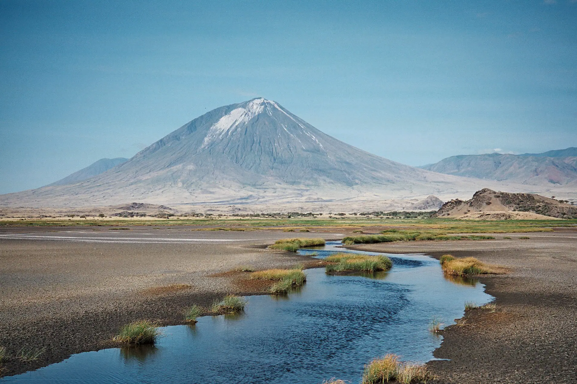 De met as bestreepte kegel van Ol Doinyo Lengai die oprijst achter de ondiepe sodavlaktes en geulen van Lake Natron
